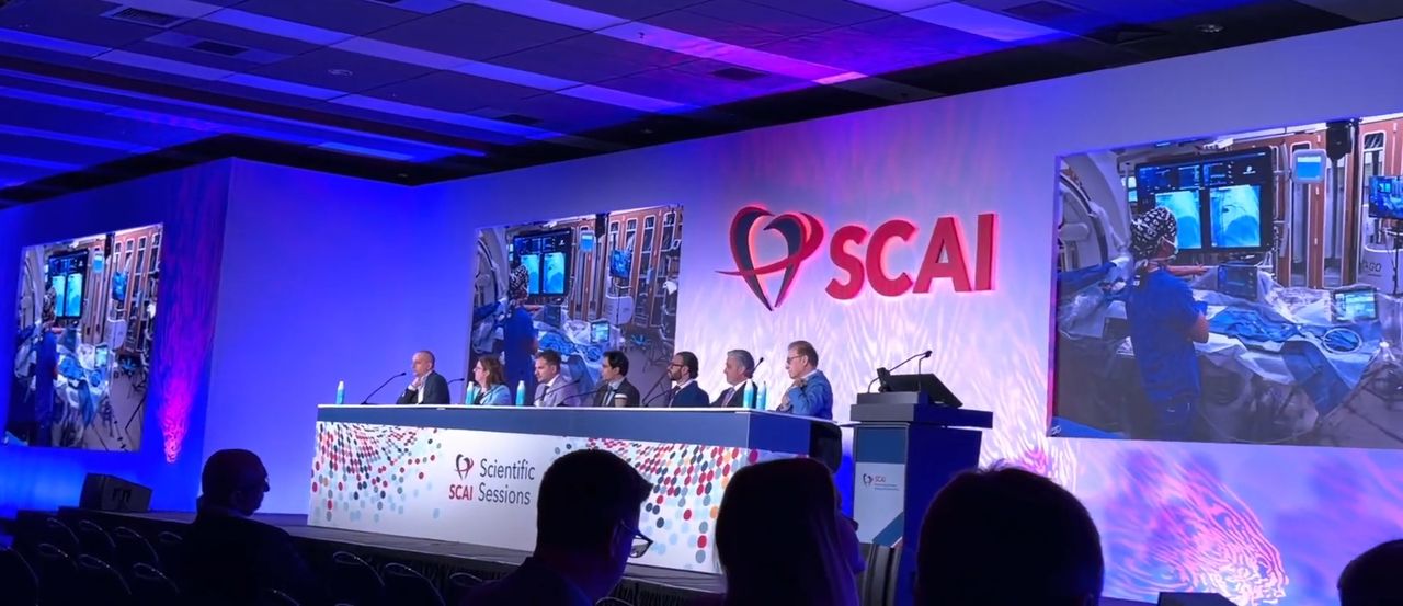 The SCAI 2026 Scientific Sessions stage: a panel of physicians beneath the SCAI sign, a live case from the cath lab on the screens behind them, and the audience in silhouette.